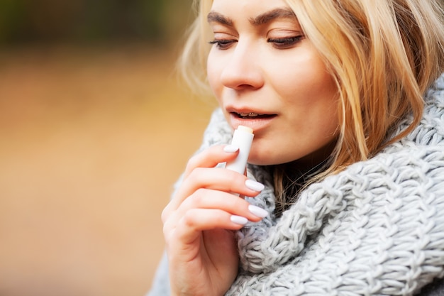 Young woman in a gray coat use hygienic lipstick in the autumn park