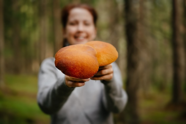 A young woman in a gray sweater in the forest holds out wild freshly picked mushrooms to the camera