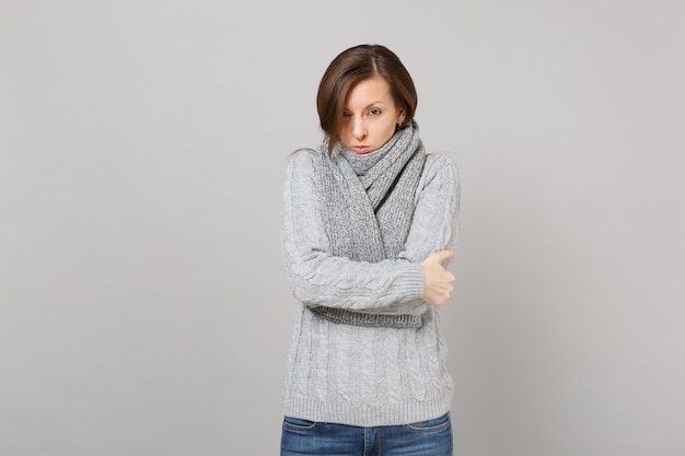 Young woman in gray sweater, scarf feeling cold hold hands folded isolated on grey wall background in studio. Healthy fashion lifestyle people sincere emotions cold season concept. Mock up copy space.