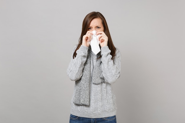 Photo young woman in gray sweater, scarf having runny nose, blowing nose to napkin isolated on grey wall background. healthy lifestyle, ill sick disease treatment, cold season concept. mock up copy space.