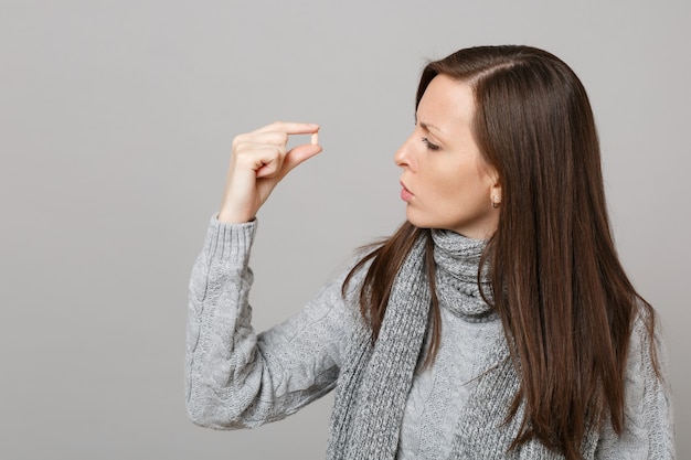 Young woman in gray sweater, scarf holding, looking on medication tablet, aspirin pill in hand isolated on grey background in studio. Healthy lifestyle ill sick disease treatment cold season concept.