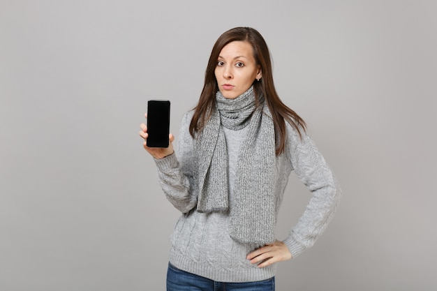 Young woman in gray sweater, scarf holding mobile phone with blank empty screen isolated on grey background. Healthy fashion lifestyle people sincere emotions, cold season concept. Mock up copy space.