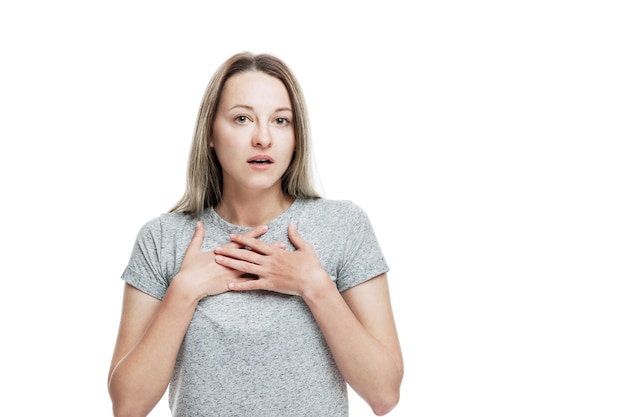 Una giovane donna con una maglietta grigia è sorpresa di aprire la bocca e di stringere le mani al petto. emozione sincera. isolato su un muro bianco