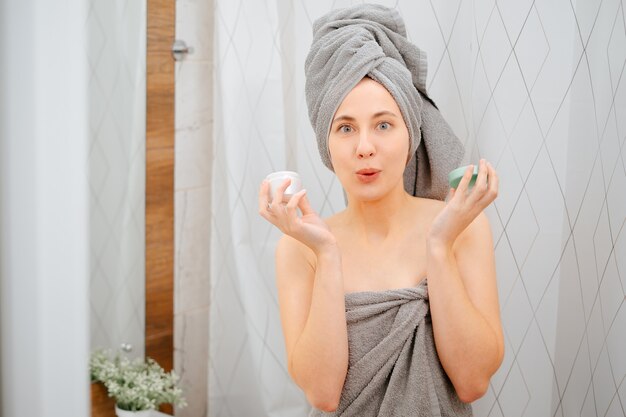 Young woman in a gray towel on the background of the bathroom holds a jar of serum for wrinkles