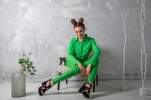 Young woman in green suit on chair Smiling girl with hairstyle and makeup poses in studio on gray background