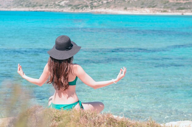 Young woman in green swimsuit and black hat practices yoga on the beach of the Mediterranean sea.