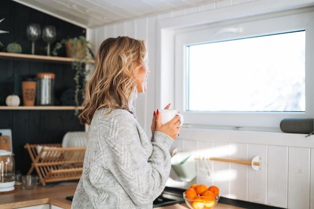 Foto giovane donna in maglione grigio con una tazza di bevanda calda in mano guardando la finestra sulla cucina a casa