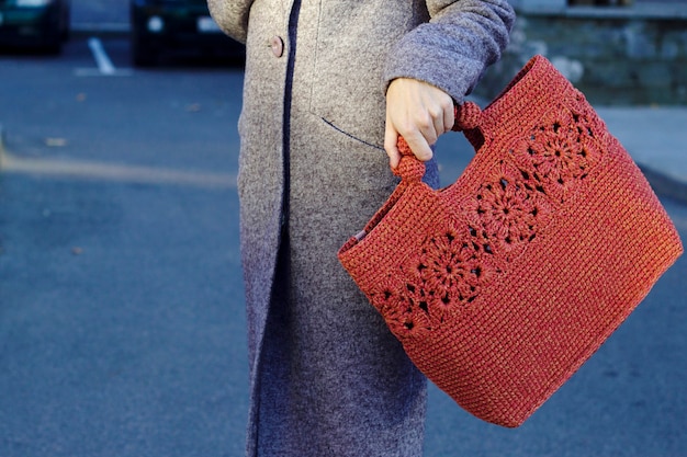 La giovane donna in cappotto caldo grigio sta andando sulla via della città con lo stile di vita della città della borsa lavorata a maglia rossa