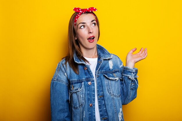 Young woman in a hair band, in a denim jacket, looks surprised to the side on a yellow surface