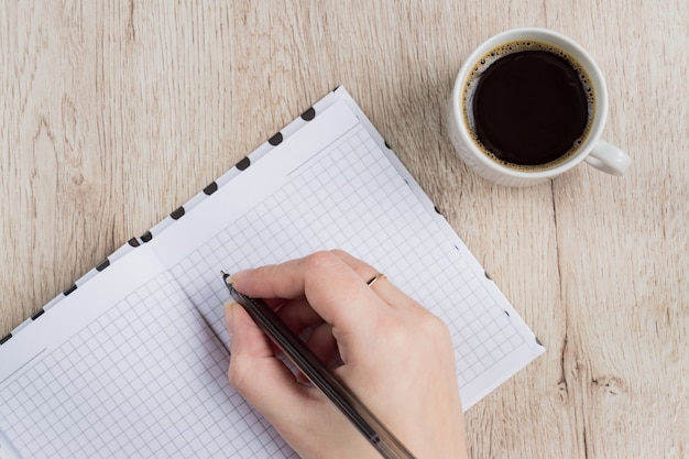 若い女性の手を保持する木製のテーブルの上のコーヒーカップの横にある黒いペンで開いているノートページ。上面図。