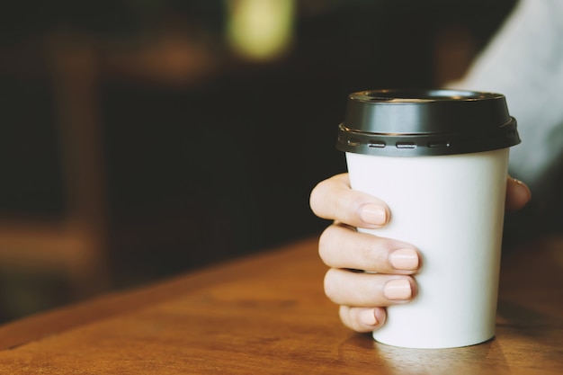 カフェのコーヒーショップで木製のテーブルで熱いコーヒーを飲みながら紙コップを持っている若い女性の手