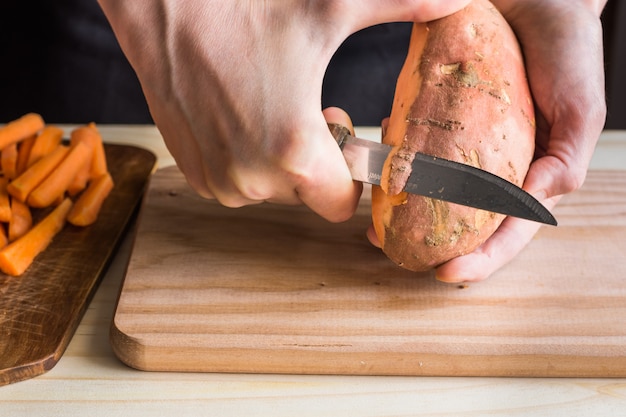 Photo young woman hands peeling with knife sweet potato over wooden cutting board