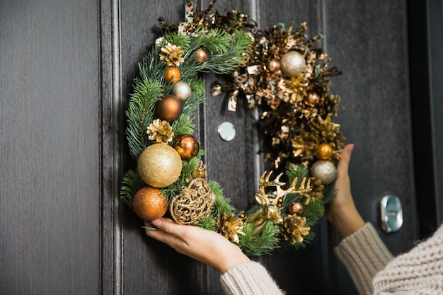 家のドアにクリスマスのお祝いの花輪をぶら下げている若い女性。冬の休日の伝統的な家の装飾、戸口にモミの木の手作りの花輪を保持している女性の手のクローズアップ。