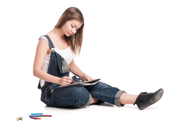 Young woman happily sitting on the floor drawing in her note pad  On a white background