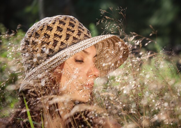 Giovane donna con un cappello tra l'erba illuminata dai raggi del sole su uno sfondo di foresta