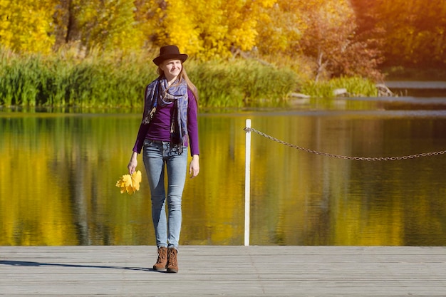 Young woman in the hat is standing on the pier Autumn sunny day Back view