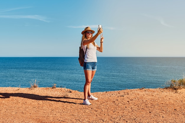 Young woman in a hat making a selfie in front of the coast in summer