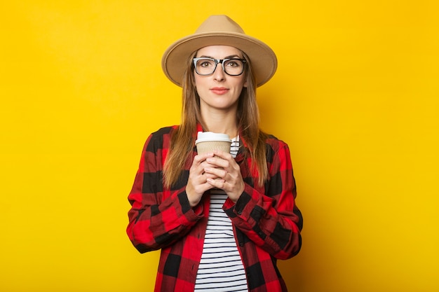 Young woman in hat and plaid shirt holding paper cup with coffee on yellow