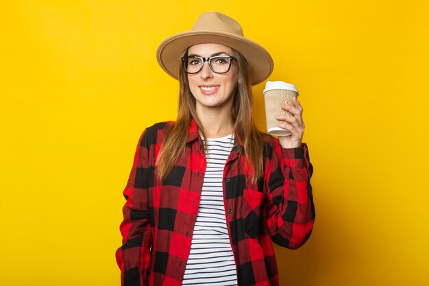 Young woman in hat and plaid shirt holding paper cup with coffee on yellow