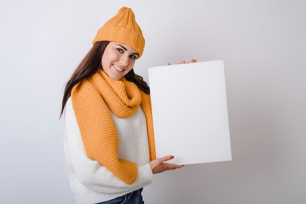 Young woman in a hat and scarf holding a white sheet in her hands on a gray background