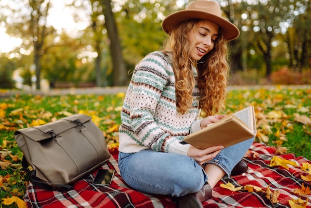 Young woman in hat sits on a plaid in autumn park and reads a book Concept of nature education