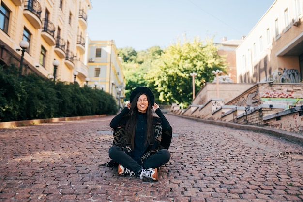 Young woman in hat sitting at city on cobblestone