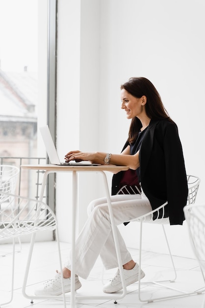 Young woman have a break watching online videos and trainings\
on laptop preparing for conference cheerful girl with laptop typing\
text and chatting with friends and family in white cafe