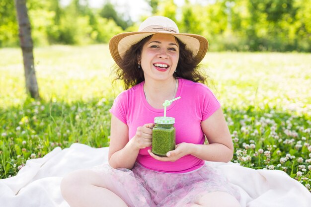 若い女性は公園で楽しんだり、ピクニックで緑のスムージーを飲みます。