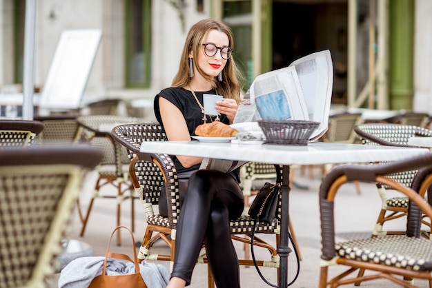 Young woman having a breakfast with coffee and croissant reading newspaper outdoors at the typical french cafe terrace in France