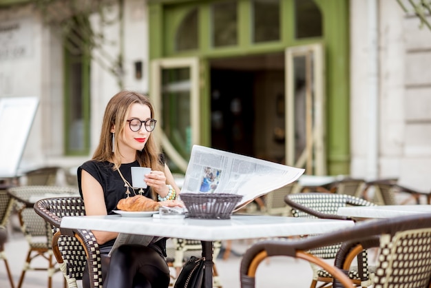フランスの典型的なフレンチカフェテラスで屋外で新聞を読んでコーヒーとクロワッサンと一緒に朝食をとっている若い女性