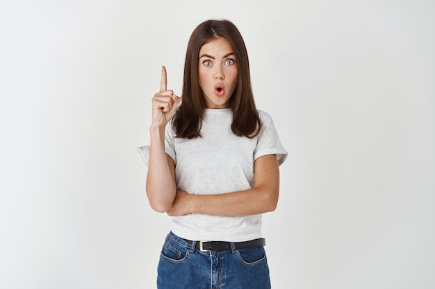 Young woman having an idea, raising finger and saying suggestion, have plan, standing over white wall