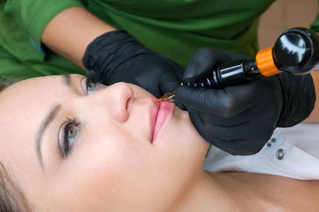 Young woman having permanent makeup on her lips at the beauticians salon. Natural green background