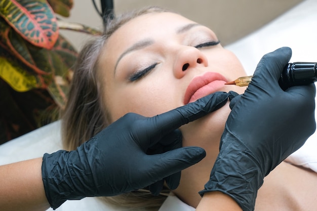 Young woman having permanent makeup on her lips at the beauticians salon. Natural green background