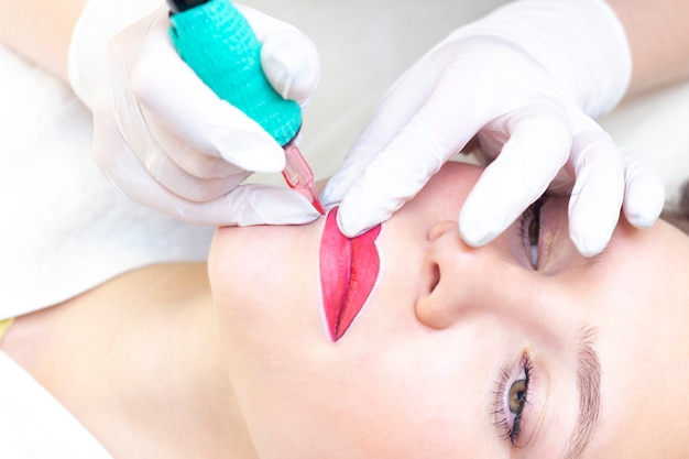 Young woman having permanent makeup on her lips at the beauticians salon. Permanent Makeup drawing a contour with a white lip pencil