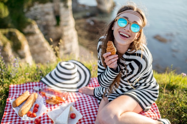 フランスの岩の多い海岸線に座ってワイン、チーズ、パンとピクニックをしている若い女性