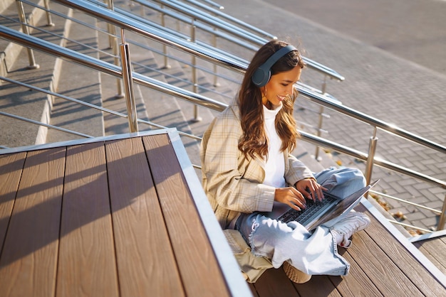 Young woman in headphone with laptop on city street Online training remote work in social networks