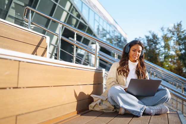 Young woman in headphone with laptop on city street Online training remote work in social networks