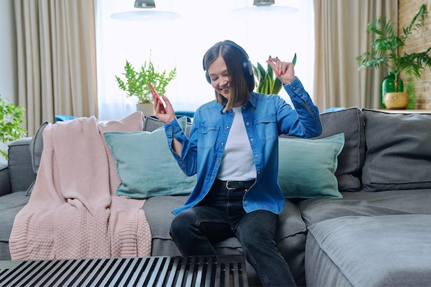 Young woman in headphones enjoying music dancing sitting on sofa at home