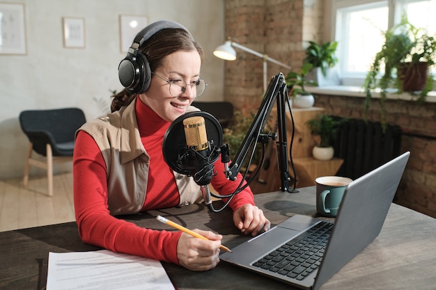 Young woman in headphones sitting at the table working on laptop and recording podcast speaking in microphone