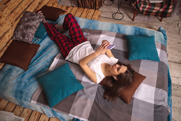 A young woman in headphones and with a mobile phone in her hands lies on the bed and takes a selfie. The girl takes pictures of herself on the phone and smiles.