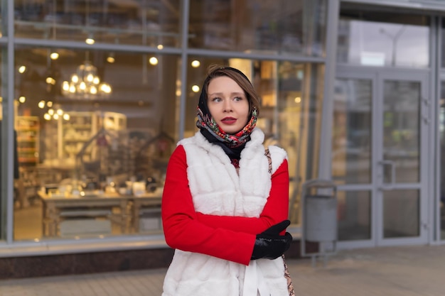 Young woman in a headscarf in cold weather stands outside against the background of a blurry bright shop window