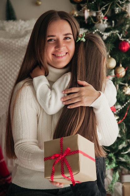 Young woman and her daughter sitting in front of the Christmas tree and holding a present