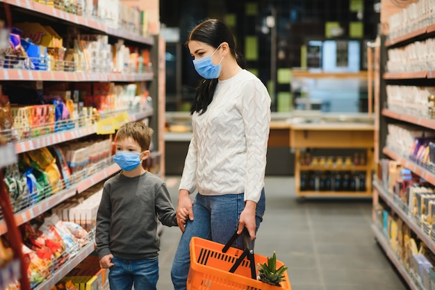 若い女性と保護用マスクを身に着けている彼女の子供はスーパーマーケットで食べ物を買う