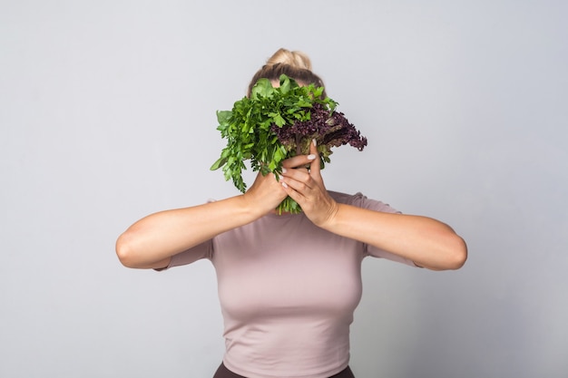 Giovane donna che nasconde il viso dietro un mazzo di erbe, verdure a foglia verde, con in mano lattuga acetosella e sorridente guardando la fotocamera, alimentazione sana, cibo biologico. foto in studio, sfondo grigio