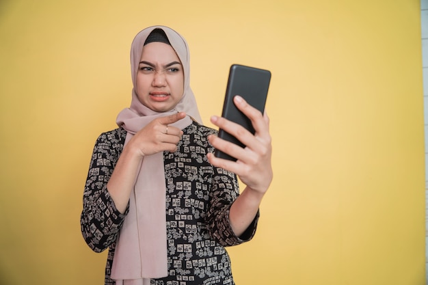 Young woman in hijab disgusted while looking at smartphone screen with hand pointing gesture