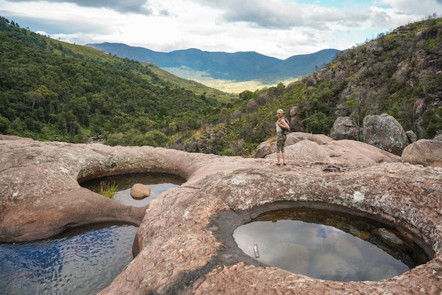 Young woman in hiking clothes standing on stones near natural water pool blurred trees and mountains background view form behind typical scenery seen during trek in Andringitra Madagascar