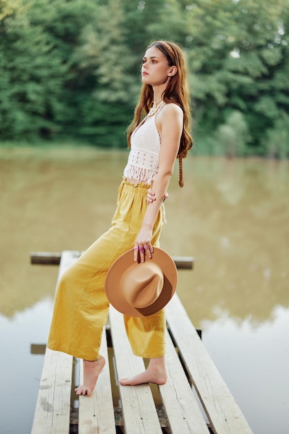 A young woman in a hippie look and ecodress dancing outdoors by the lake wearing a hat and yellow pants in the summer sunset