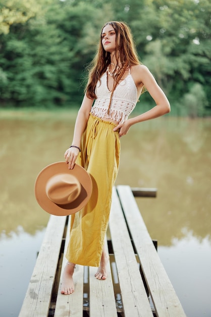 A young woman in a hippie look and ecodress dancing outdoors by the lake wearing a hat and yellow pants in the summer sunset