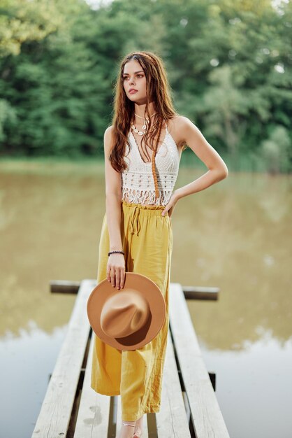 A young woman in a hippie look and ecodress dancing outdoors by the lake wearing a hat and yellow pants in the summer sunset