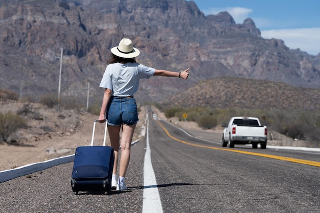 Young woman hitchhiking on a scenic highway road with blue baggage bag